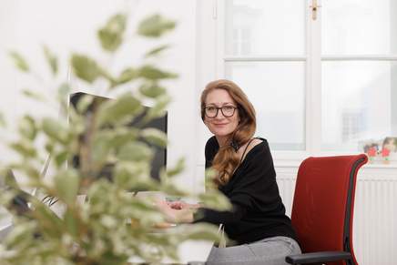 Cristina sitzt in ihren Büro an ihren Schreibtisch und schaut in die Kamera. Das Foto wurde mit einem Tiefenschärfe-Effekt aufgenommen. Im Vordergrund ist unscharf ein eine Dekopflanze zu sehen.