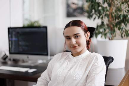 Mitarbeiterin Portrait: Frau mit roten, zusammengebundenen Haaren. Computerbildschirm steht im Hintergrund.