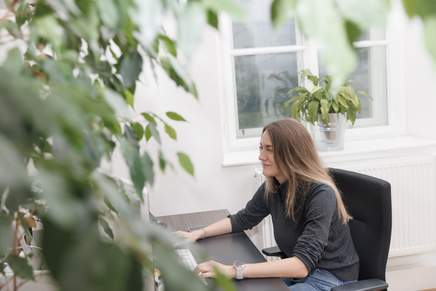 Eva sitzt an ihrem Schreibtisch und tippt etwas in den Computer ein. Im Vordergrund ist ein Pflanze unscharf ins Bild eingebaut - Tiefenschärfeeffekt.